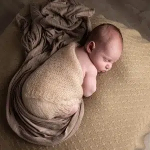durée séance photo nouveau-né Bébé endormi enveloppé dans une laine beige sur un canapé, cocooning et paisible, prêt pour une séance photo nouveau-né.