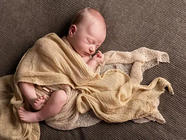 accessoires pour shooting photo bébé Bébé endormi enveloppé dans des langes beige, posé sur un fond sombre, douceur et chaleur naturelle.