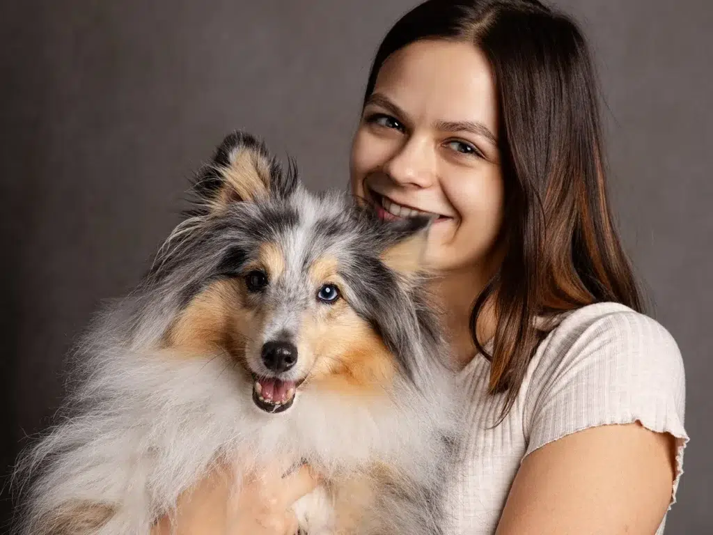 Une femme souriante pose avec son chien de race colley, tous deux près d’un fond neutre.