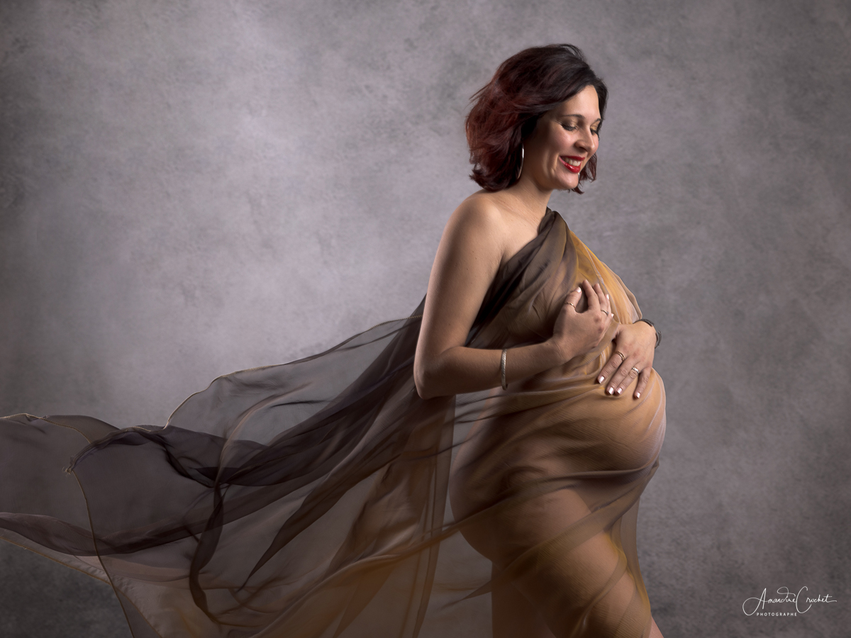 Portrait de femme enceinte enveloppée d’un voile translucide, pose élégante et sourire, ventre mis en valeur, fond neutre. idée photo grossesse mois par moi s? (intégration naturelle du mot-clé)