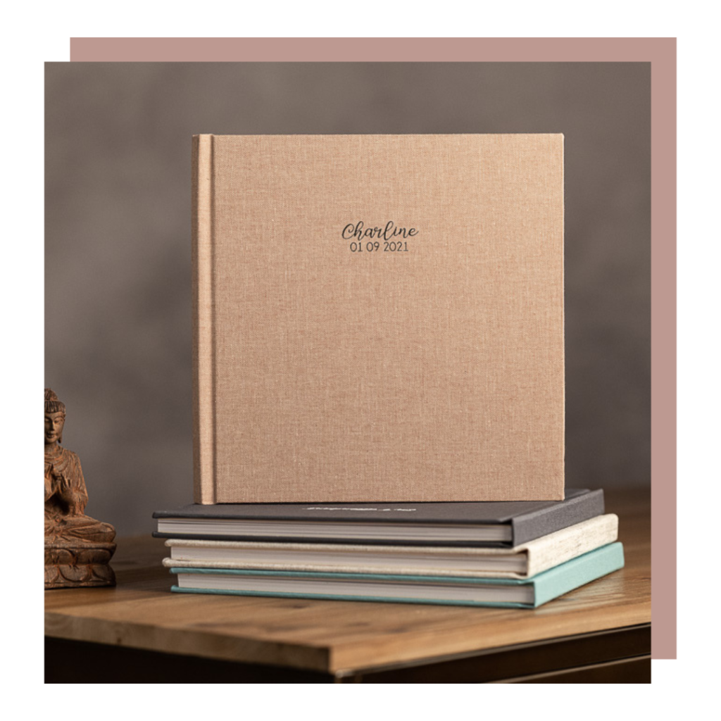 Album ou livre photo en carton beige posé sur des livres, avec le nom "Charline" et la date imprimés dessus.